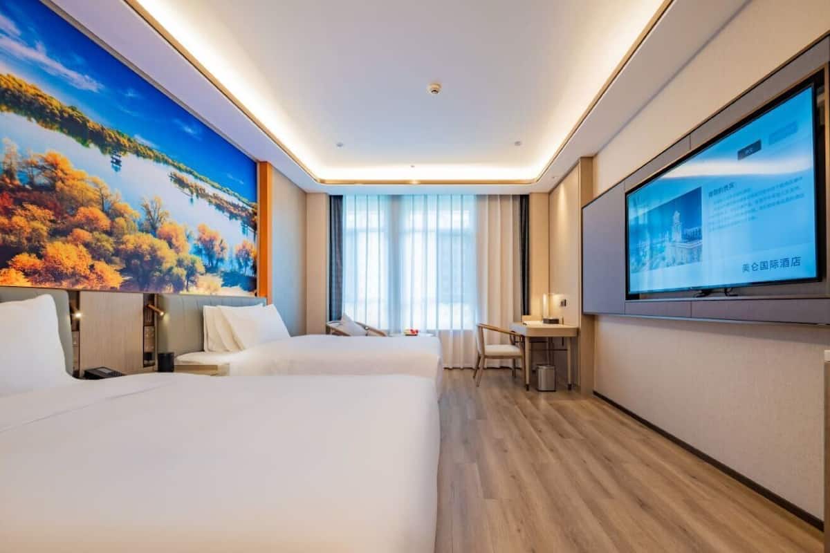Habitación de hotel con dos camas, un escritorio, una silla, un televisor y un ventanal con cortinas.