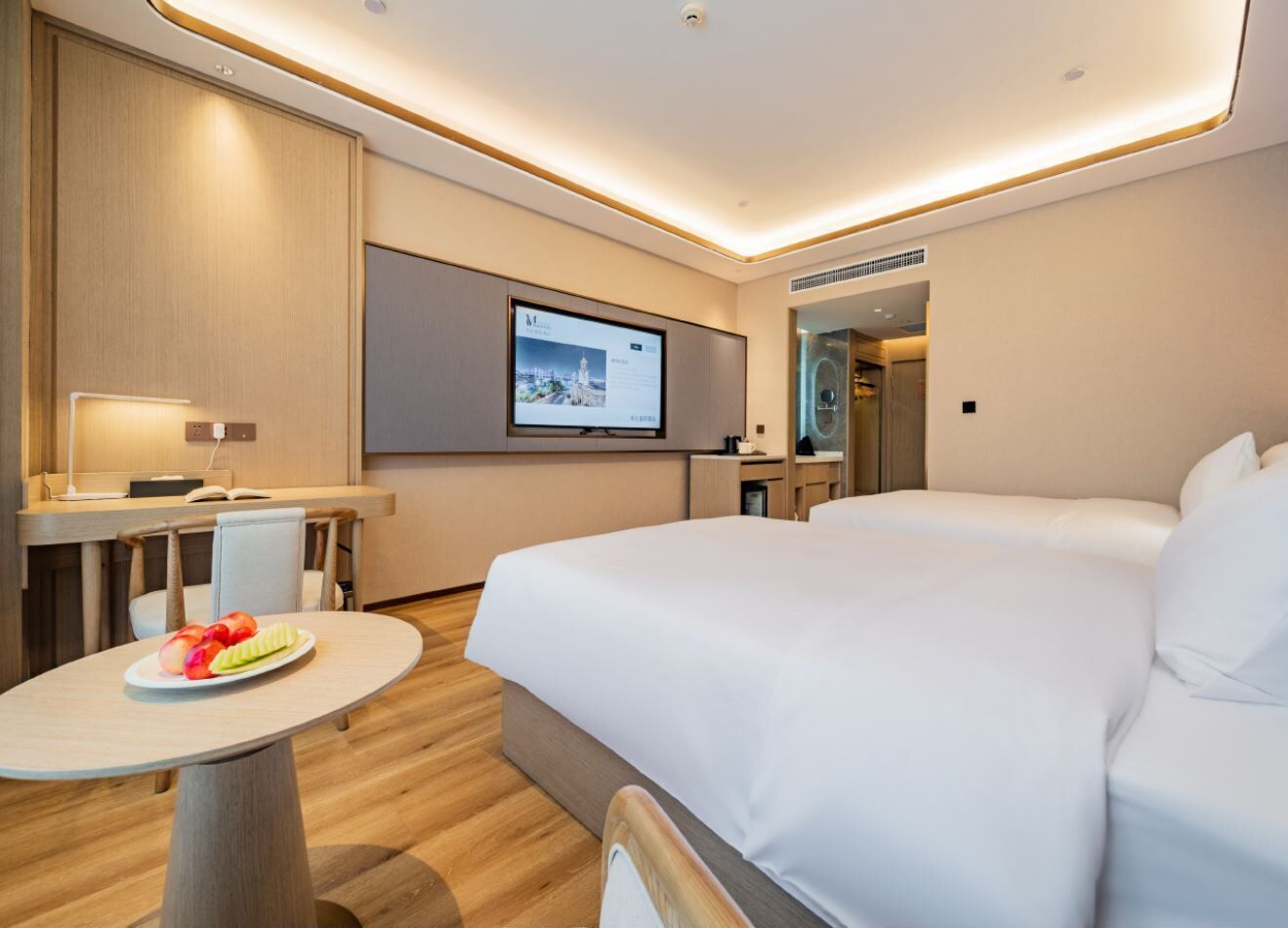 A modern hotel room with a bed, a desk, a chair, a small table with fruit, and a TV screen.