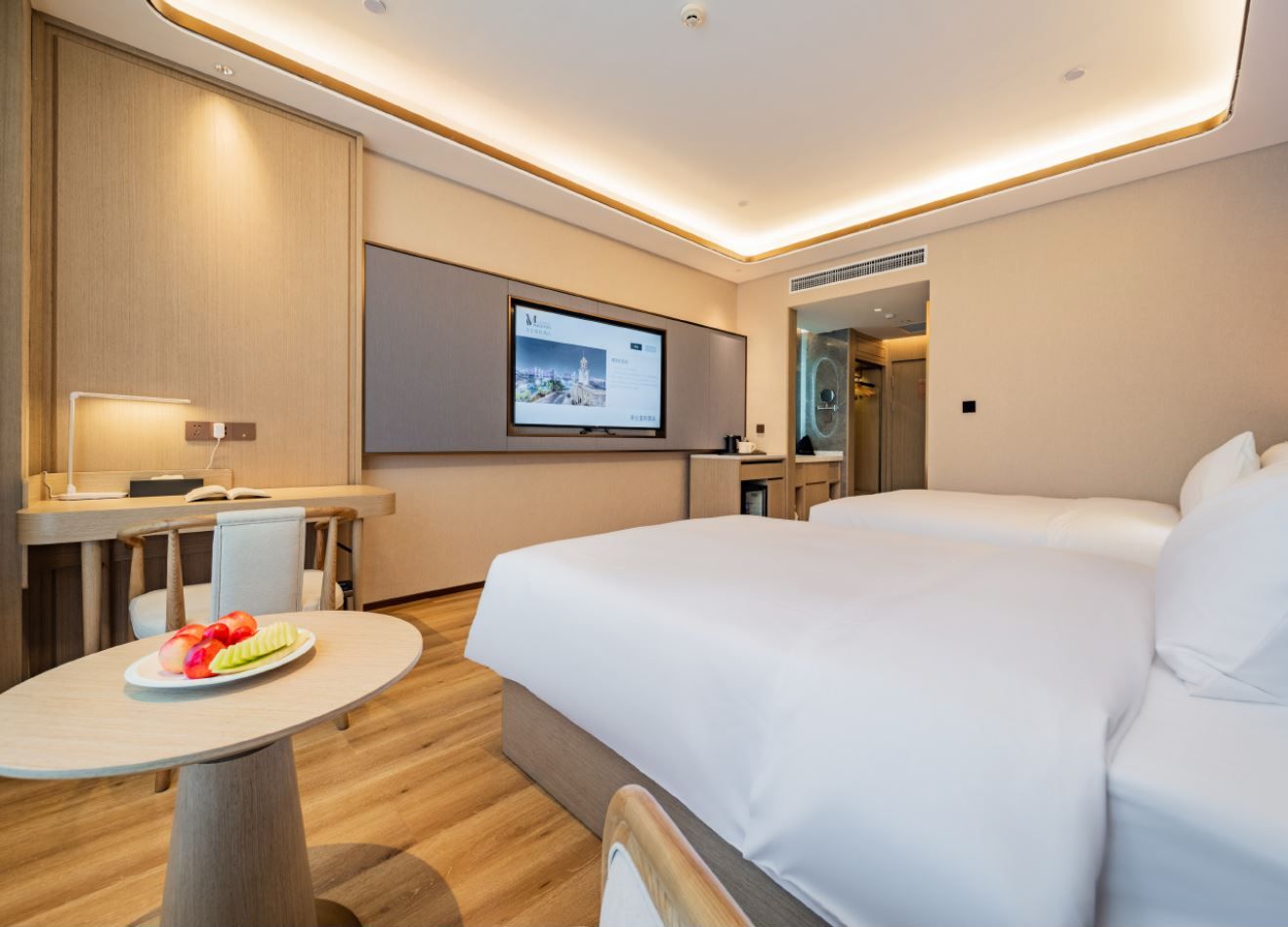 Una habitación de hotel moderna con cama, escritorio, silla, una mesita con fruta y una pantalla de televisión.