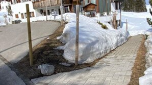 En asfalterad gångväg med snö på båda sidor, som leder fram till en byggnad med en träveranda och ett snötäckt tak.