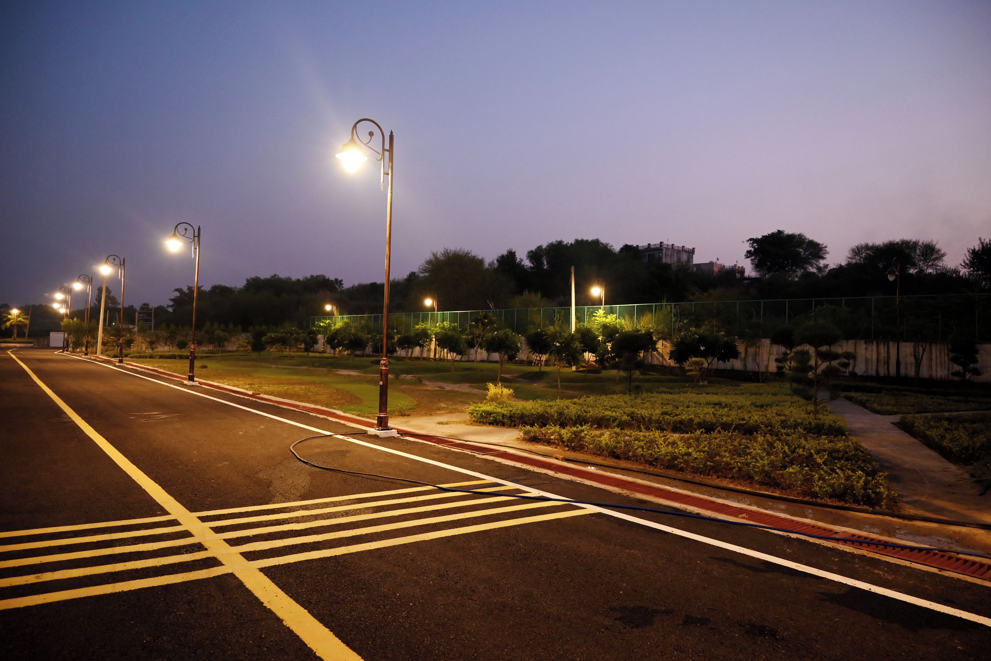 A well-lit street at dusk with clear road markings, street lamps, and a green fence.