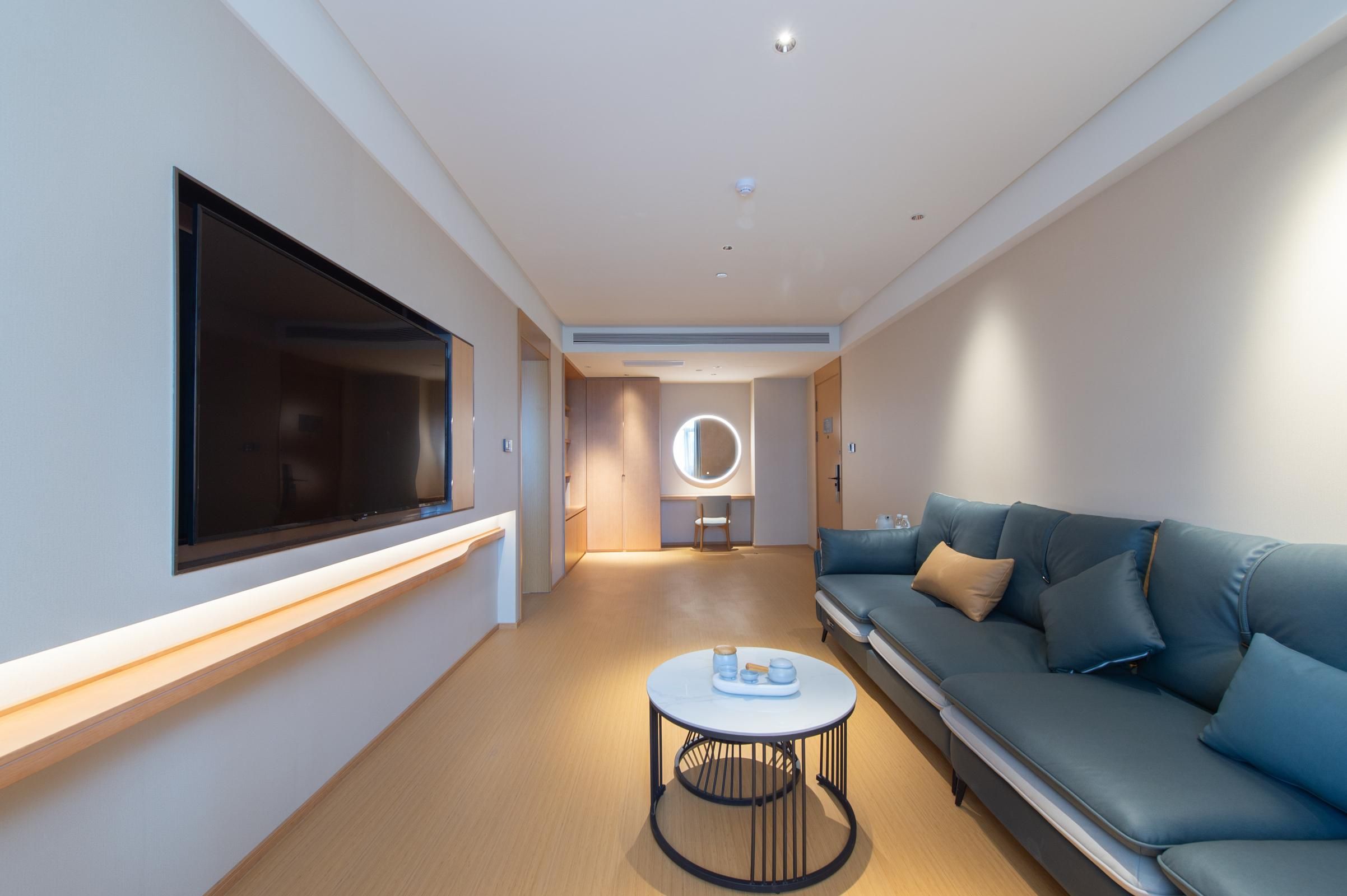 A modern hotel room with a large flat-screen TV mounted on the wall, a sofa, a round coffee table, and a circular mirror on the wall.