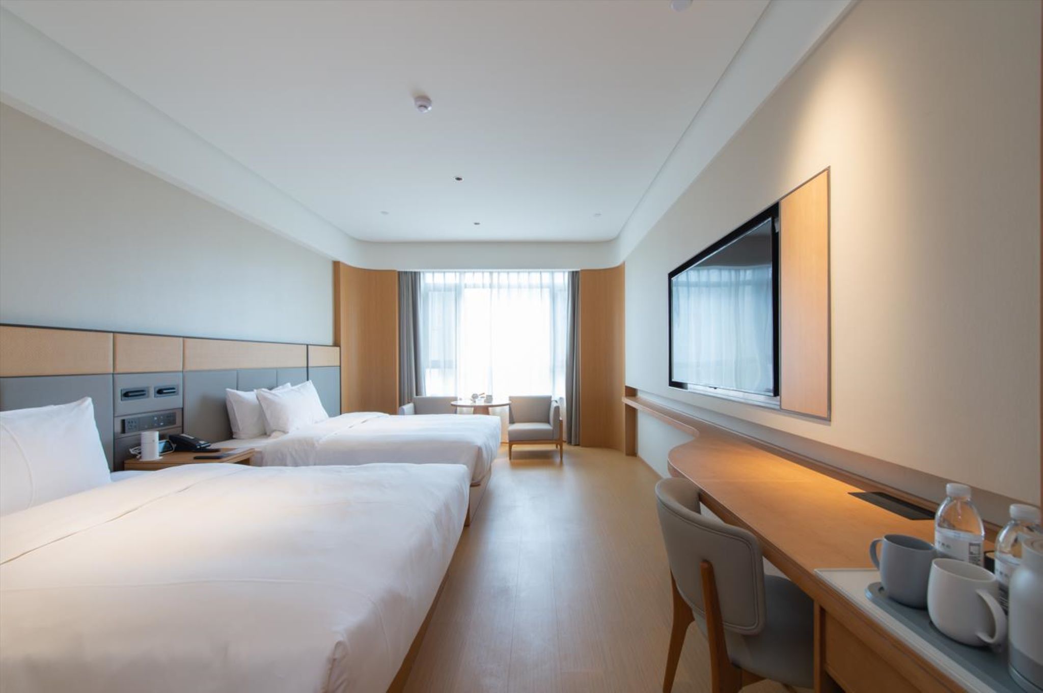 A hotel room with two beds, a desk, a chair, a TV, and a window with curtains.