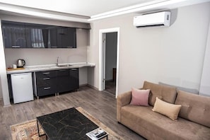 Una cocina moderna con armarios oscuros, una encimera blanca y un refrigerador pequeño. Hay un sofá color beige con cojines y una mesa de centro con una revista.