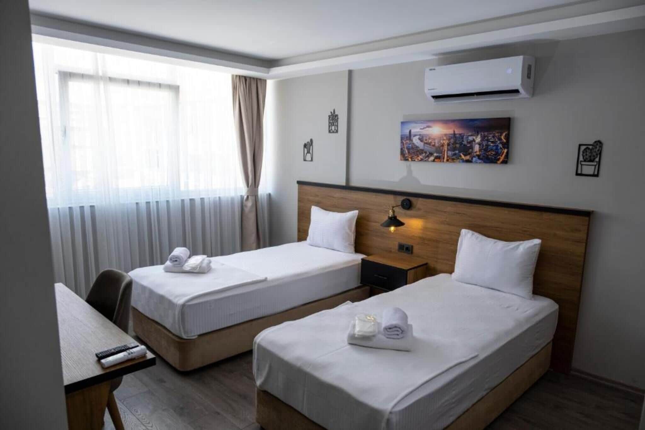 A hotel room with two beds, a desk, a window with curtains, an air conditioner, and a cityscape painting on the wall.