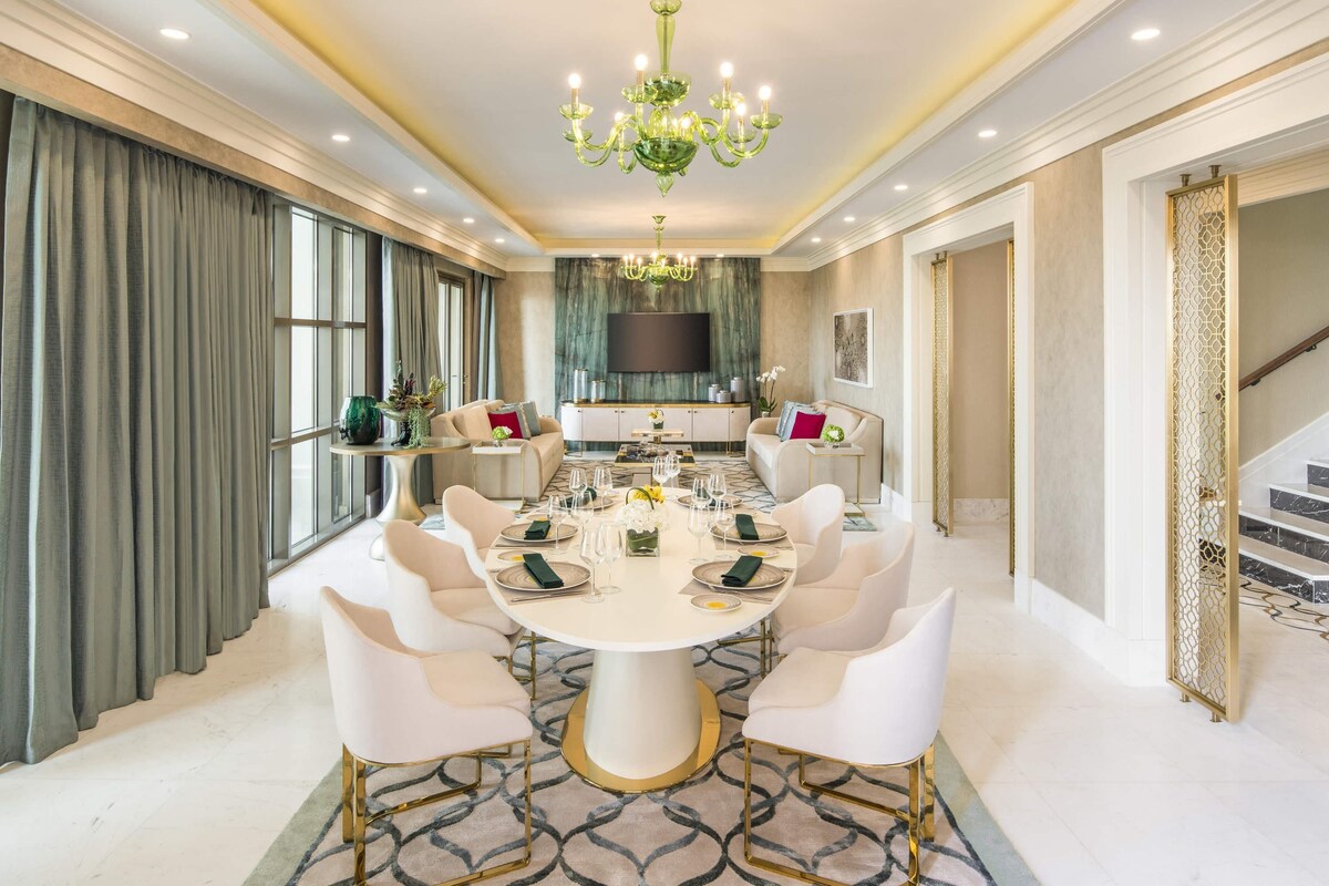 A dining room with a round table, white chairs, a chandelier, and a TV in the background.