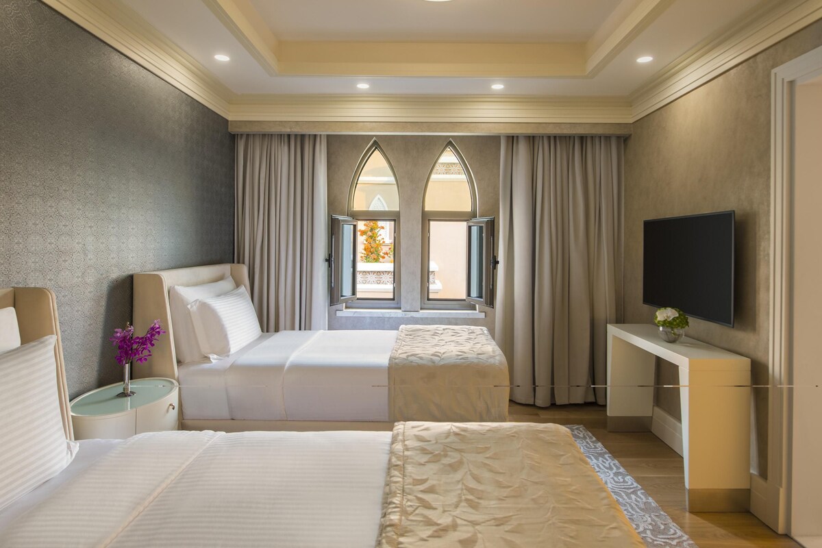 A hotel room with two beds, a TV, a small table with a vase, and a window with curtains.