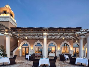 An outdoor dining area with tables and chairs, covered by a pergola, and a staircase leading to an upper level.