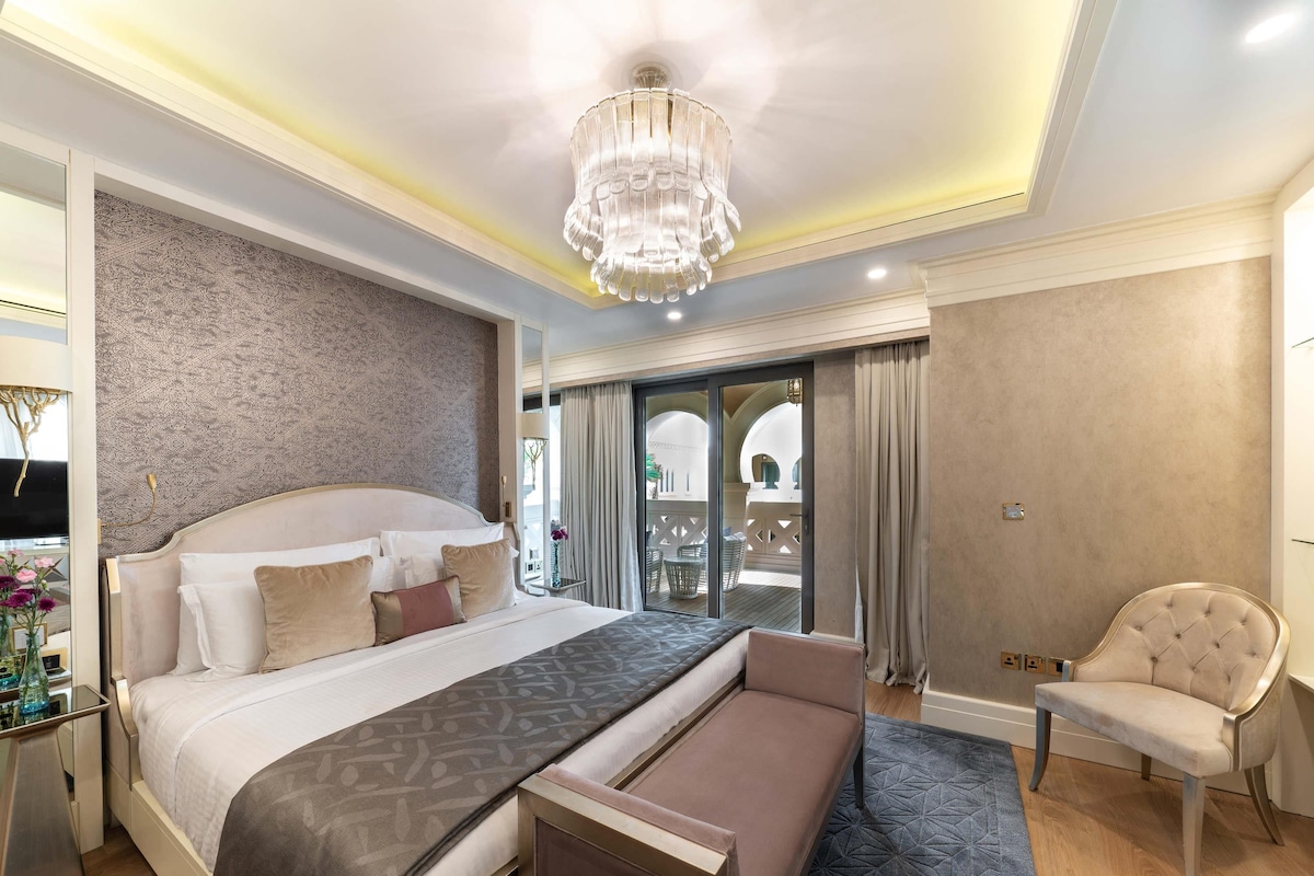 A spacious bedroom with a large bed, a chandelier, a balcony with a view, and a seating area.