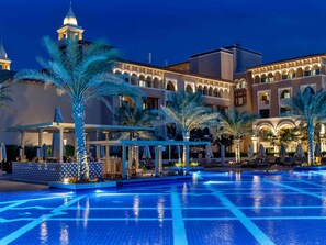 A hotel with a large swimming pool, palm trees, and a building with arches and a tower.