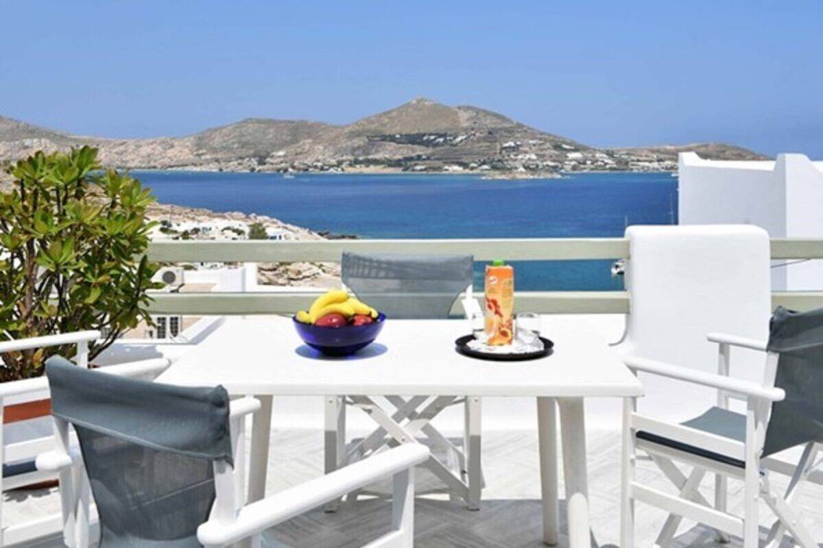 A white table with a bowl of fruit and a bottle, overlooking a coastal landscape with mountains.