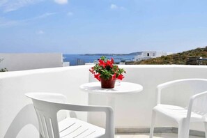 A terrace with white chairs and a table, a potted plant, and a view of the sea and buildings. - Hara Studios (Paros)