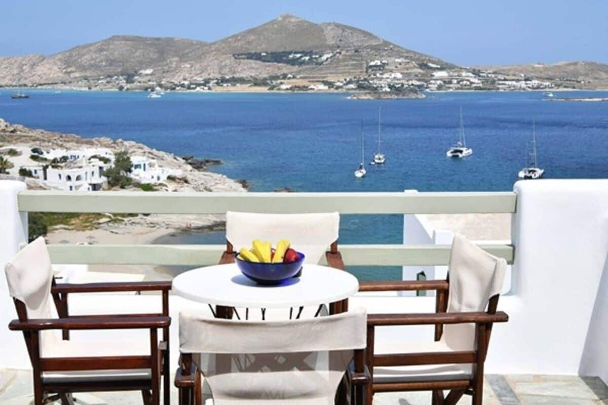 A balcony with two chairs and a table, a bowl of fruit, and a view of the sea with boats and a mountain in the background.