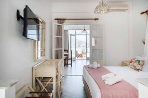 A bedroom with a bed, a desk, a chair, a TV mounted on the wall, and an open door leading to a balcony. - Hara Studios (Paros)