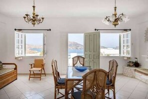 A room with a dining table, chairs, a chandelier, and large windows with a view. - Hara Studios (Paros)