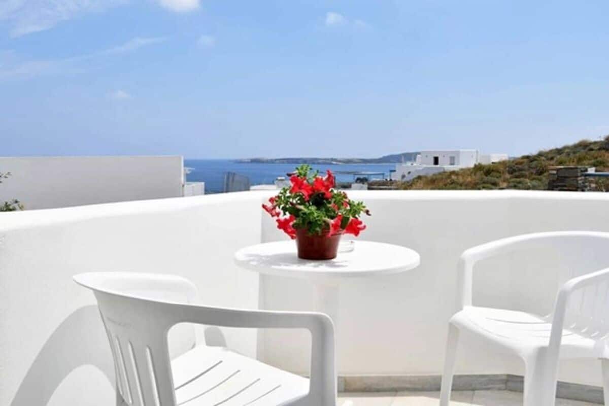 A terrace with white chairs and a table, a potted plant, and a view of the sea and buildings.