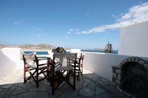 A terrace with a table and chairs, a stone fireplace, and a view of the sea and distant hills. - Hara Studios (Paros)