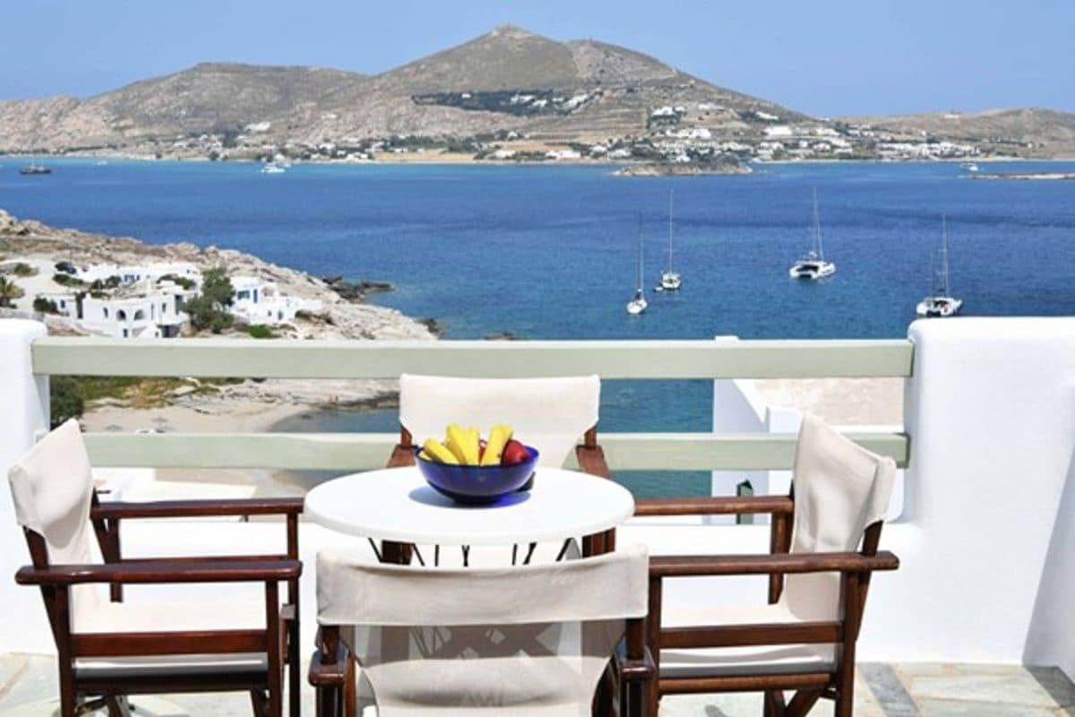 A balcony with two chairs and a table, a bowl of fruit, and a view of the sea with boats and a mountain in the background.