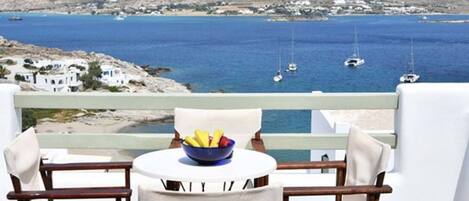 A balcony with two chairs and a table, a bowl of fruit, and a view of the sea with boats and a mountain in the background.