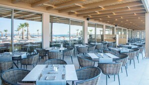 Un espacio exterior techado para comer con mesas y sillas de mimbre, y amplios ventanales con vista a la playa.