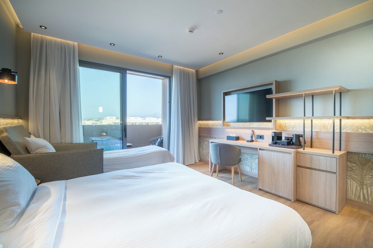 A hotel room with a large bed, a desk, a chair, a television, and a view of the city.
