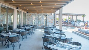 An outdoor dining area with tables and chairs, stone walls, and a poolside seating area. - Senseana Seaside Resort And Aquadventure (Analipsis)