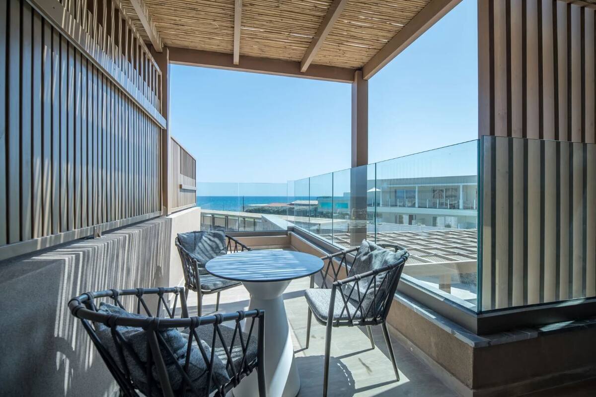 A balcony with a glass railing, two chairs, a table, and a view of the sea.