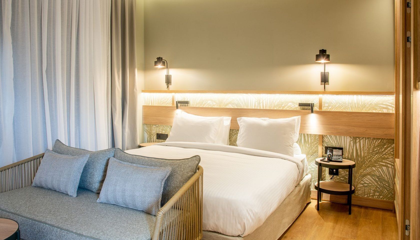 A hotel room with a bed, two bedside tables with lamps, a sofa with cushions, and a curtain.