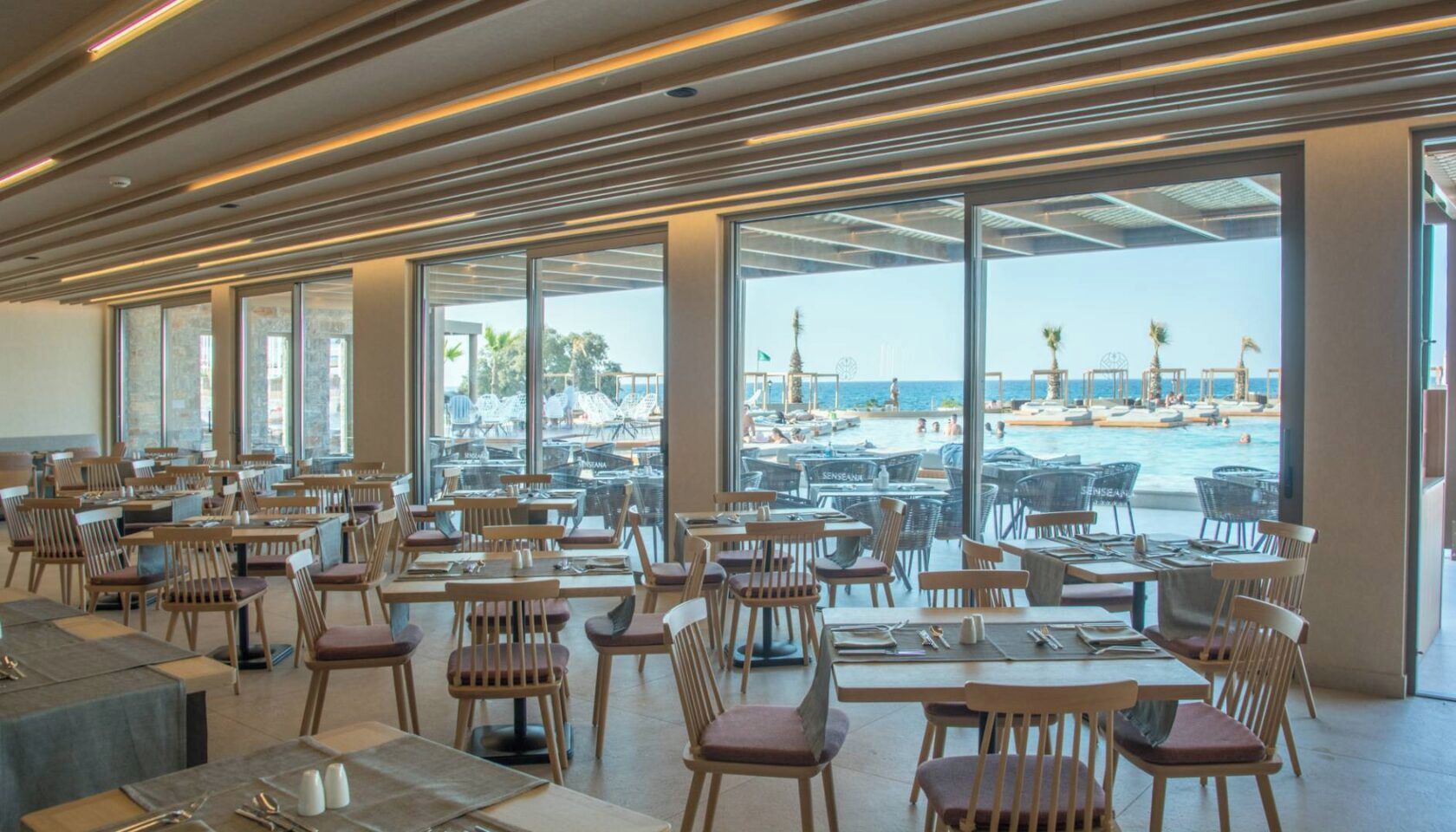 A spacious dining area with tables and chairs, large windows offering a view of a pool and beach.