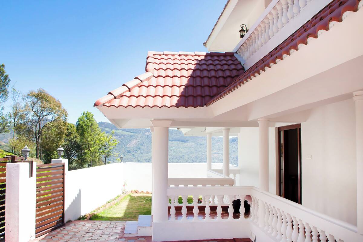 A white building with a red-tiled roof, a balcony with white balustrades, and a view of green trees and mountains in the background.