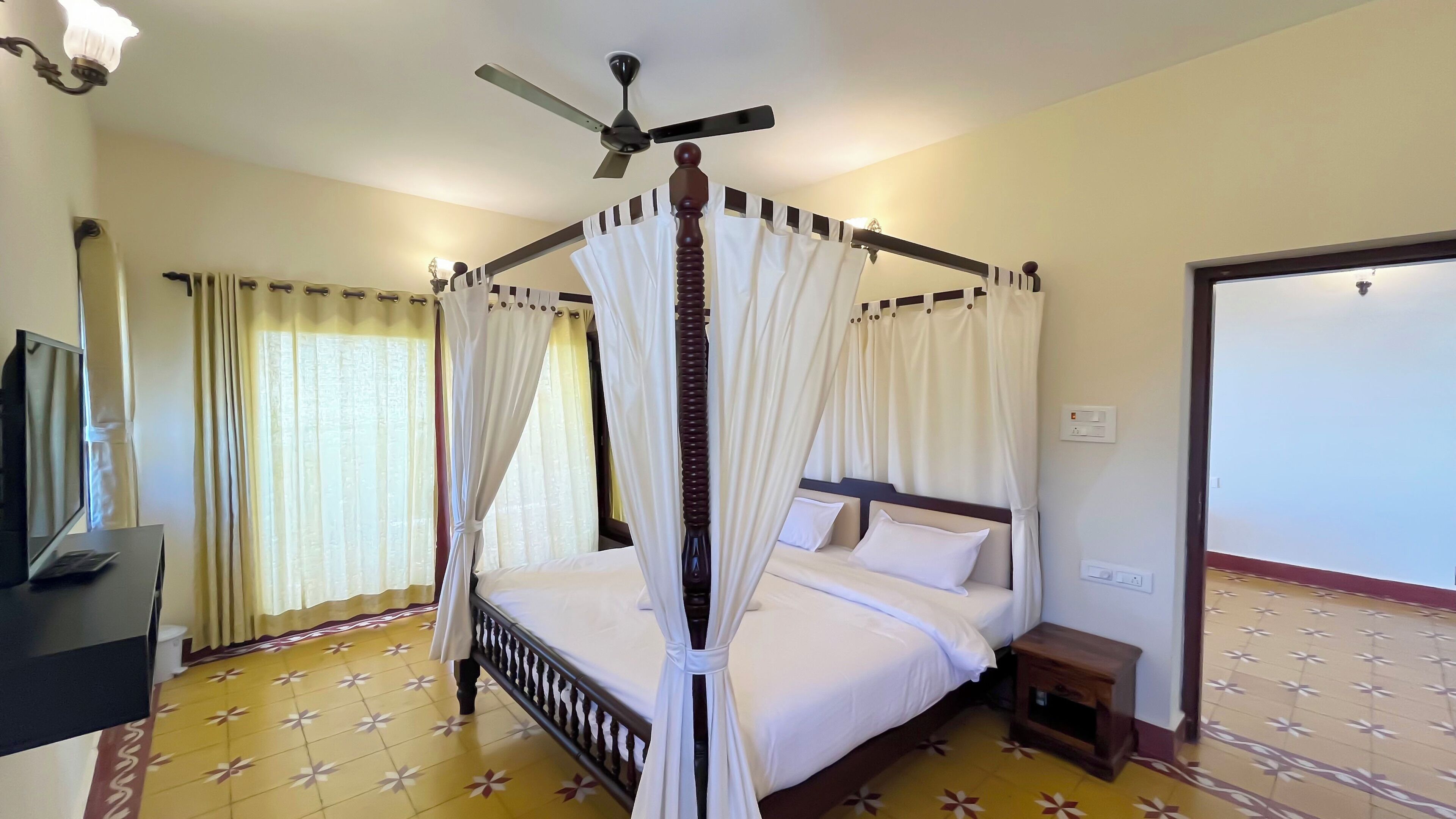 A four-poster canopy bed with white bedding, a ceiling fan, and a tiled floor.