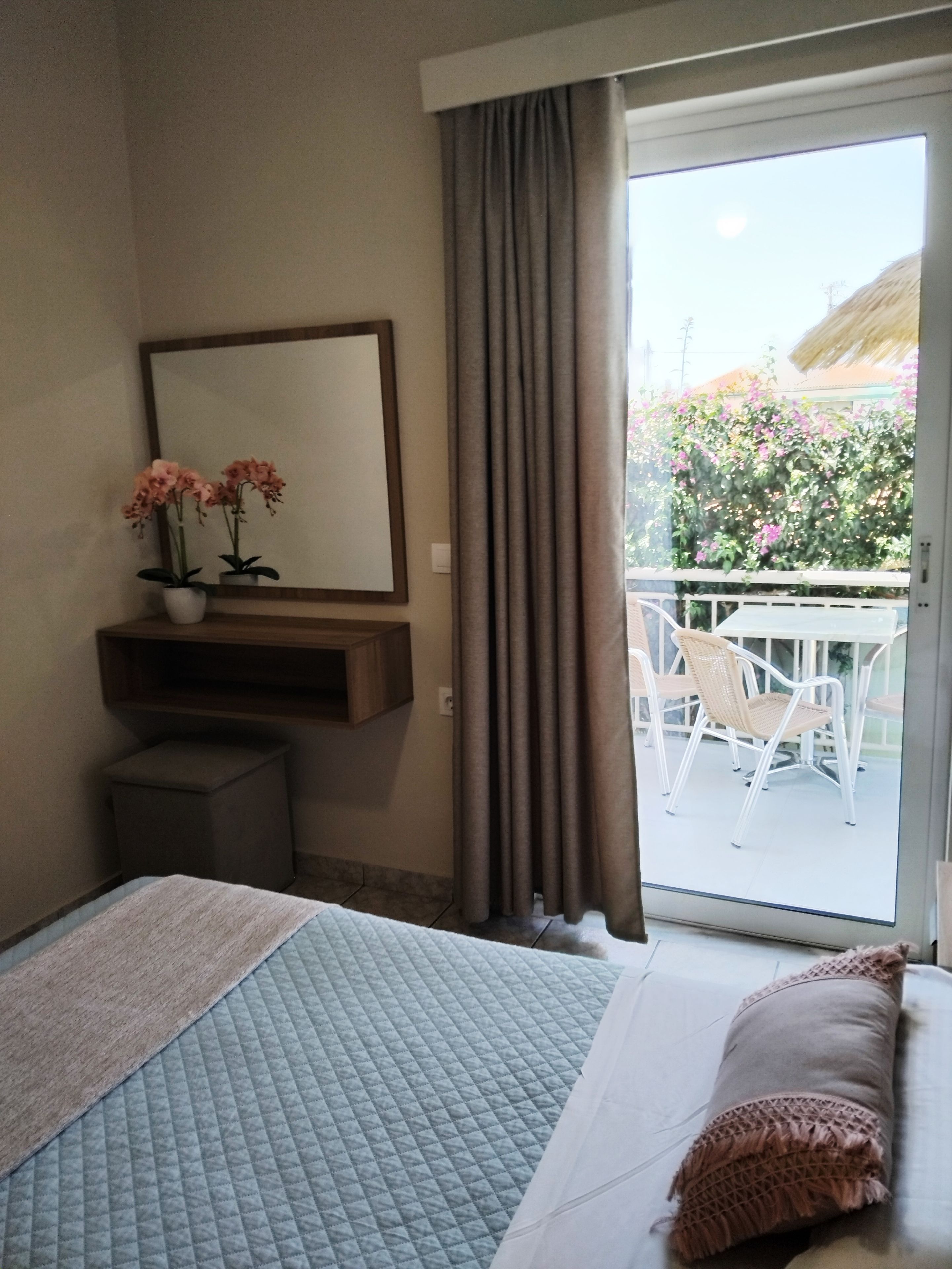 A hotel room with a bed, a bedside table with a vase of flowers, a mirror, and a view of an outdoor area with a table and chairs.