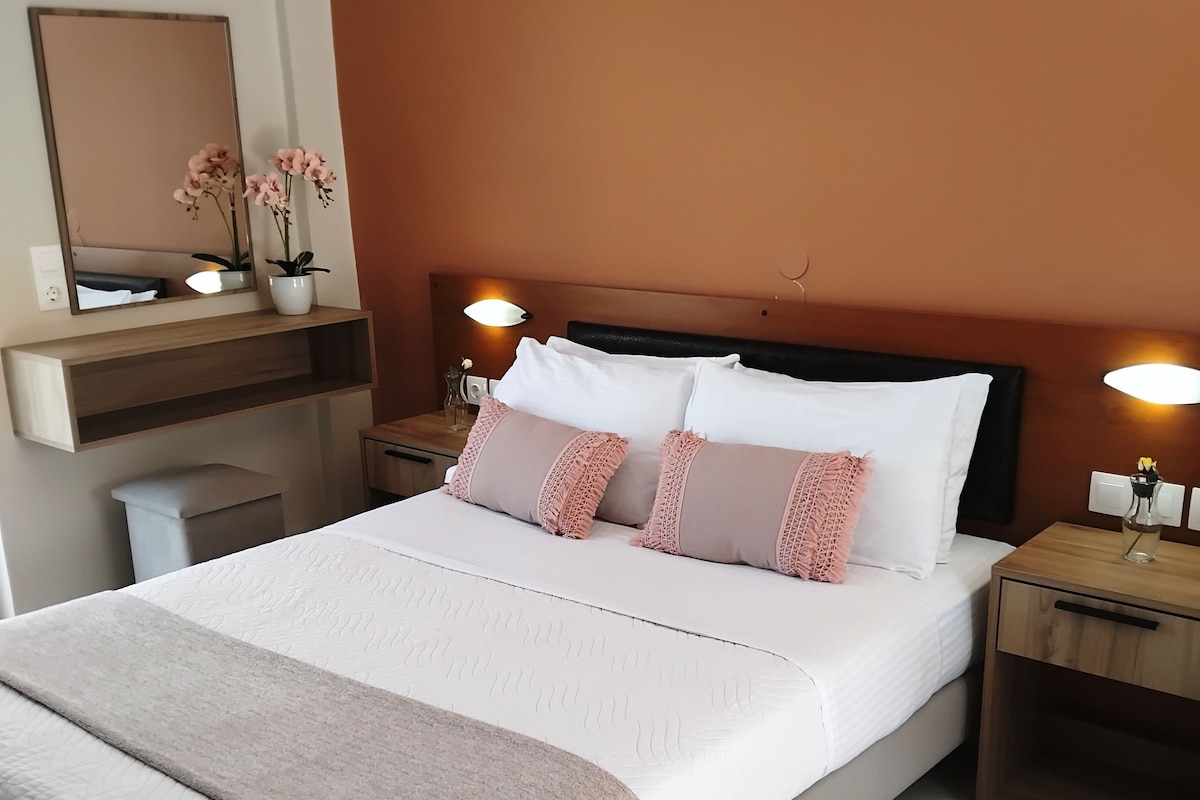 Cama bem arrumada com edredom branco e almofadas cor-de-rosa, mesa de cabeceira com abajur e espelho fixado na parede.