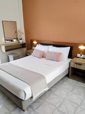 A neatly made bed with a white comforter and pillows, a bedside table with a lamp, a wall-mounted shelf with a vase, and a mirror.