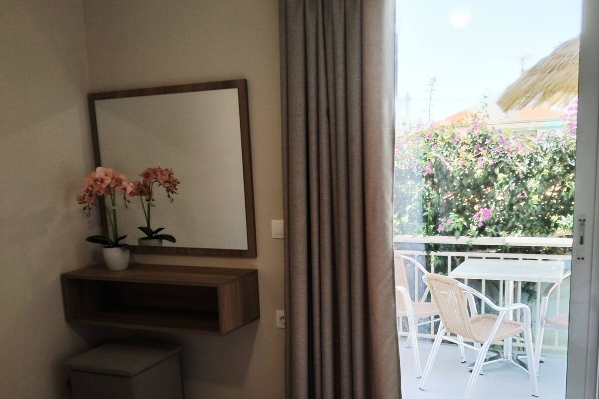 A hotel room with a bed, a bedside table with a vase of flowers, a mirror, and a view of an outdoor area with a table and chairs.
