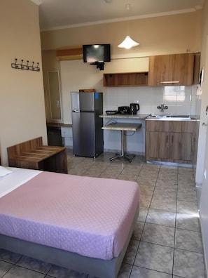 Quarto compacto com cama, cozinha compacta, pequena mesa de jantar e televisão fixada na parede.