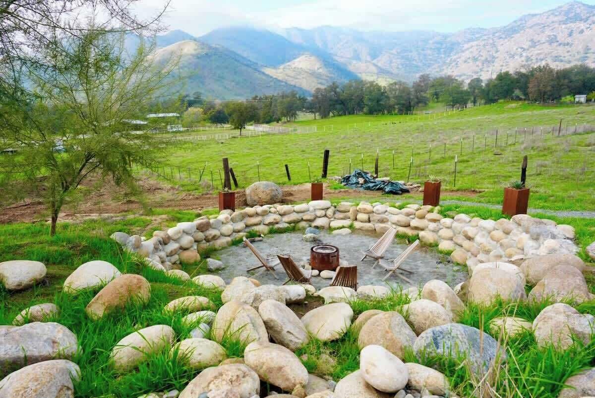Ein runder Feuerkorb aus Stein mit Stühlen, umgeben von Steinen und Gras, in einer grünen Wiese mit Bergen im Hintergrund.
