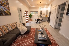 A modern living room with a sofa, coffee table, and books. There is a dining area with chairs and a table, and a kitchen with appliances. - Sequoia Park: Cozy House w/ private hot tub (Three Rivers)