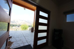 A room with a large wooden door leading to an outdoor patio with a stone floor and a mountain view.