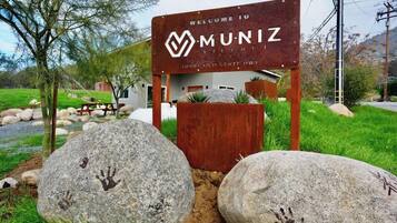 Ein Willkommensschild für Muniz Village mit einer rustikalen Holzkonstruktion, großen Felsen mit Handabdrücken und einer grünen Wiese.