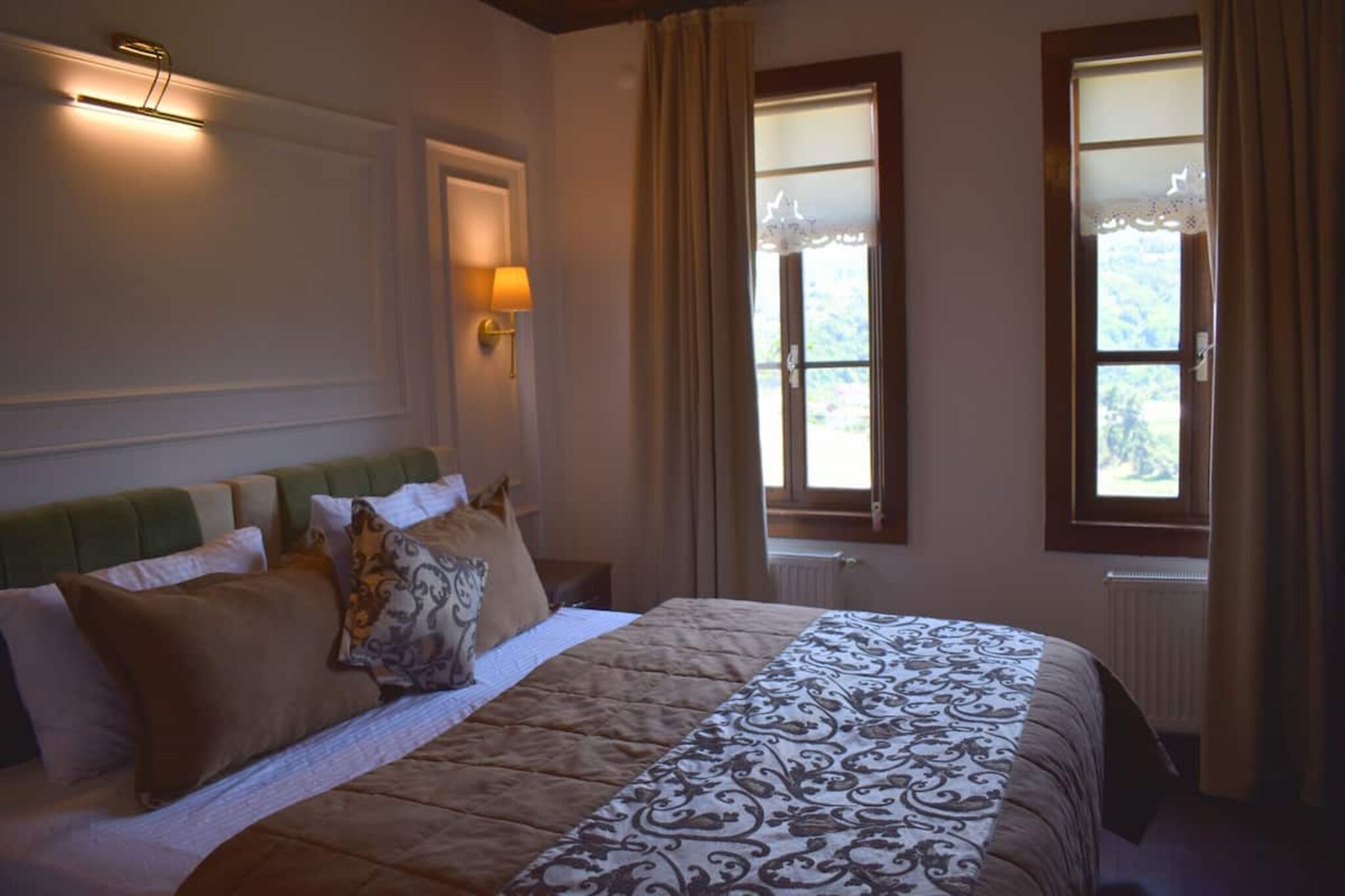 A bedroom with a bed, a wall-mounted lamp, and two windows with curtains.