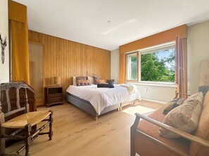 A bedroom with a large bed, wooden walls, a window with curtains, a chair, and a bedside table. - Entre Pistes & Golf - Crans Montana by Interhome (Crans-Montana)