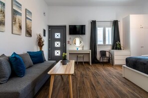 Quarto compacto com sofá, escrivaninha, cama, televisão e janela com cortinas.