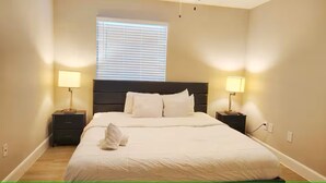 A neatly made bed with white linens, two bedside tables with lamps, a window with blinds, and a wooden floor. - Rust King 2Bed 2Bath / By TMC /5 min walk to NRG (Houston)