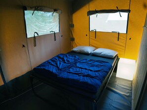 Un lit simple avec une couverture bleue, deux oreillers blancs et une fenêtre avec un rideau.