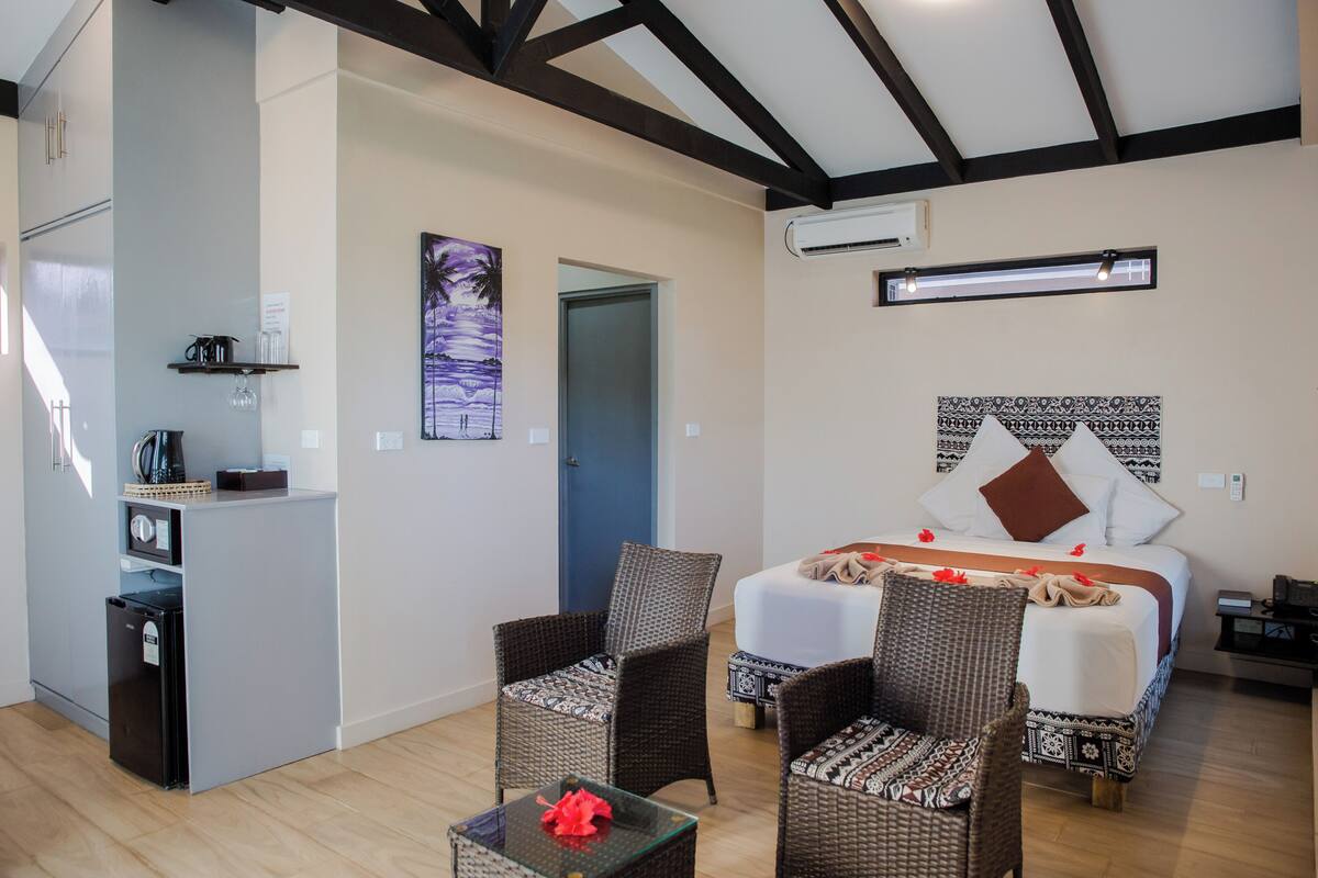 A hotel room with a bed, two wicker chairs, a small table, a kitchenette, and a painting on the wall.