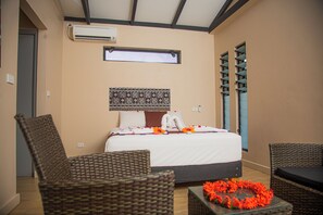 A hotel room with a bed, wicker chairs, a glass coffee table with flowers, a patterned tapestry, and a window with blinds. - Seatiki Fiji (Sigatoka)