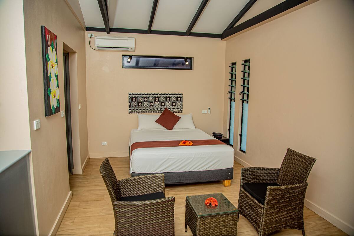 A hotel room with a bed, two wicker chairs, a small table with a flower, a wall tapestry, and an air conditioning unit.