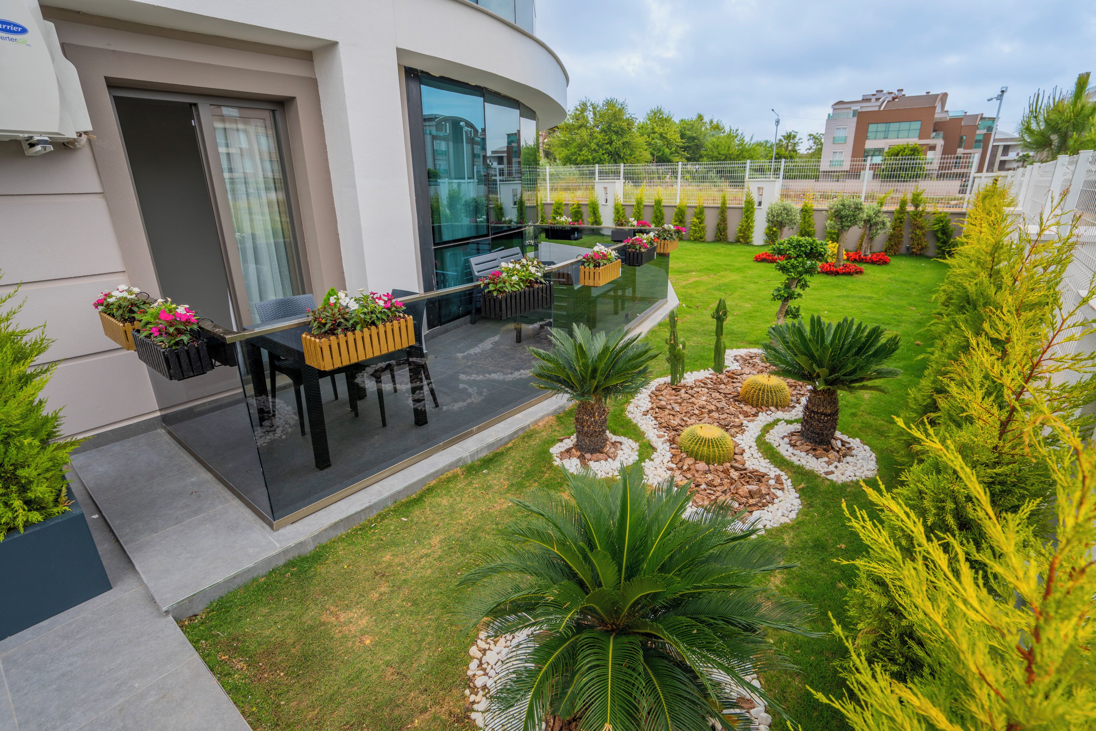 A modern outdoor patio with a glass balustrade, a seating area with a table and chairs, and a well-maintained garden with various plants and a small cactus.