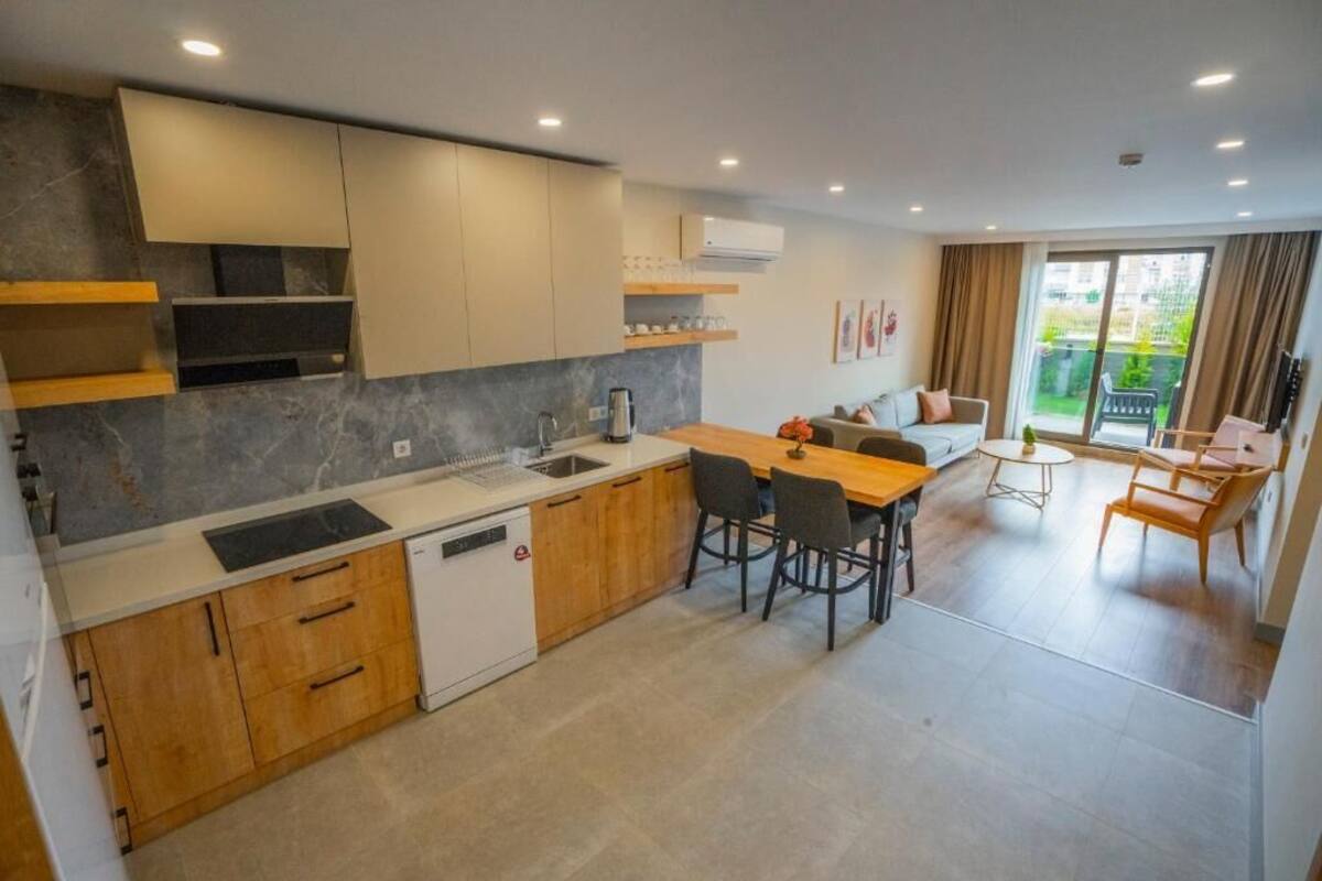 A modern kitchen with wooden cabinets, a dining area with a table and chairs, and a view of a garden through a sliding door.